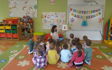 Dzieci siedzą na kolorowym dywanie i słuchają bibliotekarki oczytającej książkę