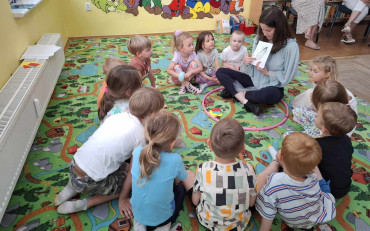 Dzieci siedzą na kolorowym dywanie, a bibliotekarka pokazuje obrazek