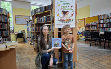 Bibliotekarka wraz z dzieckiem, nowym czytelnikiem, pokazują książkę, karte oraz...