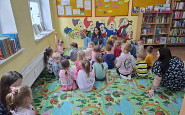 Dzieci siedzą na kolorowym dywanie i słuchają bibliotekarki, opowiadającej historie