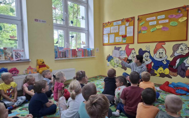 Dzieci siedzą na dywanie i słuchają opowiadania czytanego przez bibliotekarkę