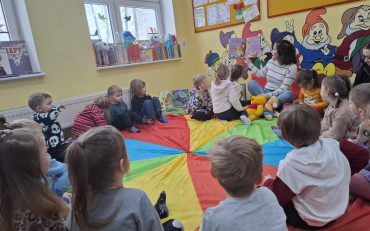 Dzieci siedzą na dywanie i słuchają opowiadania czytanego przez bibliotekarkę